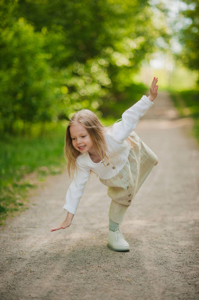A little girl in a white dress learning to stand on one leg in the Park in the summer on the path. - Fotografie, Obrázek