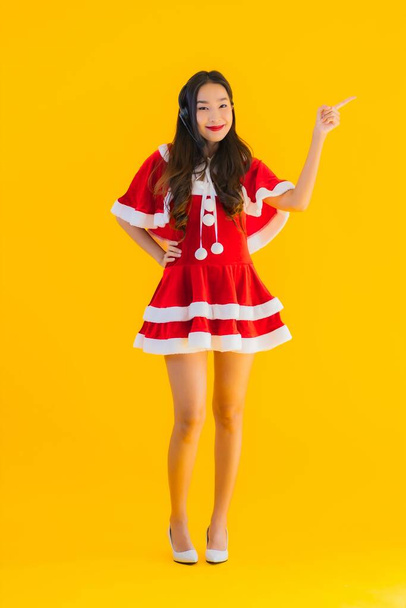 Portrét krásná mladá asijská žena nosit vánoční oblečení a klobouk s sluchátkem nebo sluchátka pro call centrum na žlutém izolovaném pozadí - Fotografie, Obrázek