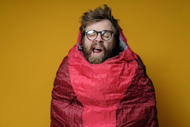 Funny, yawning man in a sleeping bag on a yellow background, he sleeps and imaginary himself in a wonderful travel. - Photo, image