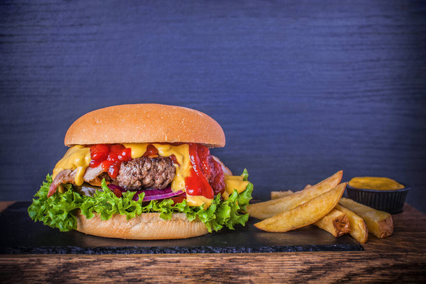 Burger with beef, bacon, cheese, fresh vegetables and sauces on plate over wooden background. For restaurant menu. Craft beef burger and french fries on a board with a stone and wooden desk - Fotoğraf, Görsel