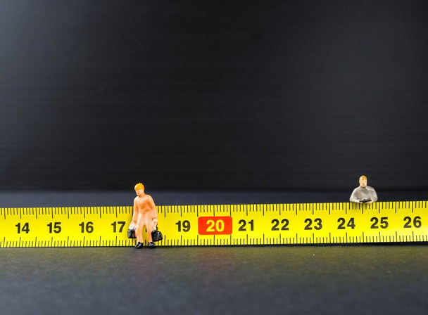 ウイルス発生を制御するための移動制御中の1メートルの社会的距離の概念。黄色い測定テープの他に2名の小型労働者が立っている。左側のフィギュアに焦点を当てる. - 写真・画像