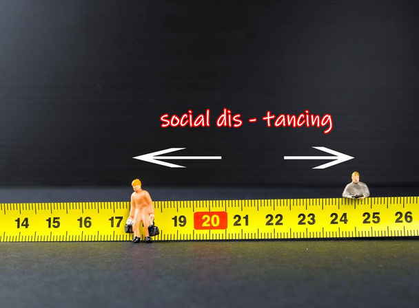 ウイルス発生を制御するための移動制御中の1メートルの社会的距離の概念。黄色い測定テープの他に2名の小型労働者が立っている。左側のフィギュアに焦点を当てる. - 写真・画像