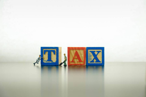 Conceptual image of working on annual tax return. Wooden block letter TAX on dark reflective surface. 2 miniature workers lifting block letter visible. Focus at block letter X. - Photo, Image