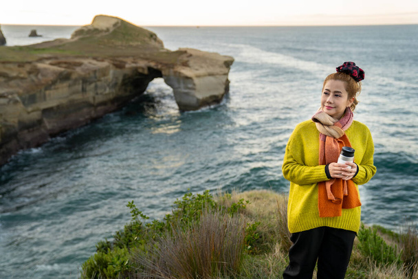 Kaunis aasialainen nainen, jolla on kahvikuppi auringonnousun aikana tunnelissa rannalla, Otago Peninsula, Uusi-Seelanti. - Valokuva, kuva