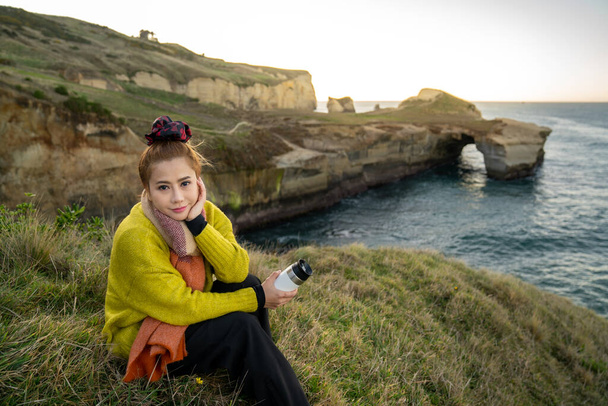 Kaunis aasialainen nainen, jolla on kahvikuppi auringonnousun aikana tunnelissa rannalla, Otago Peninsula, Uusi-Seelanti. - Valokuva, kuva