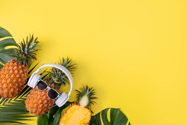 Funny pineapple wearing white headphone, listen music, isolated on yellow background with tropical palm leaves, top view, flat lay design concept. - Photo, Image
