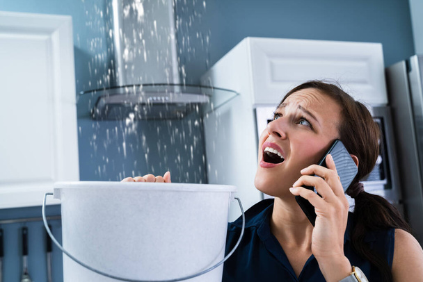 Besorgte Frau hält Eimer, während Wassertropfen aus Decke in Küche austreten - Foto, Bild