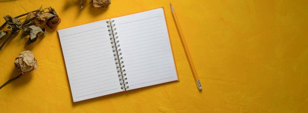 Vista superior del espacio de trabajo creativo con cuaderno en blanco abierto, lápiz, rosas secas y espacio de copia sobre fondo de hormigón amarillo
 - Foto, imagen