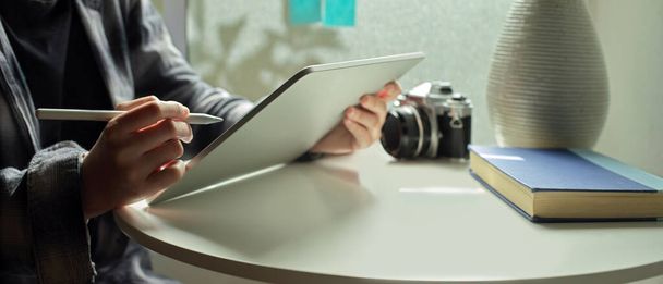 Przycięte ujęcie freelancerki pracującej z tabletem cyfrowym na stoliku z aparatem fotograficznym, książką i wazonem przy oknie  - Zdjęcie, obraz