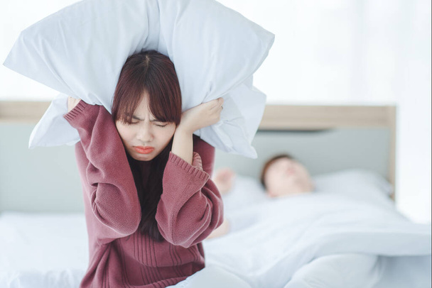 Пара в постели мужчина спит и храпит в то время как женщина не может спать закрывая уши с большими подушками, женщина блокирует уши с подушкой в то время как мужчина храпит на кровати
 - Фото, изображение