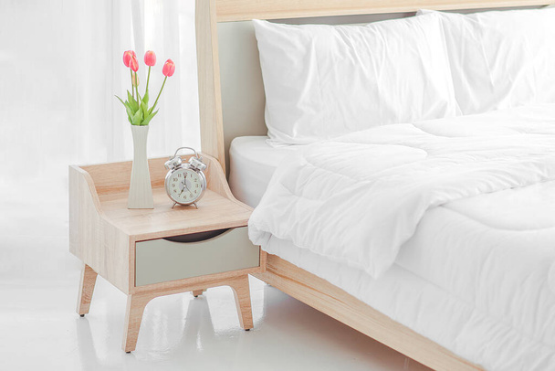 Close up of modern bed and bedside cabinet with clock and flower vase in the bedroom, bedroom. букет на тумбочке
 - Фото, изображение