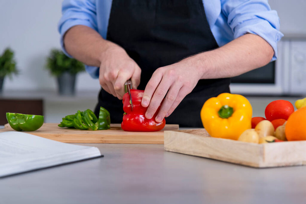Close up of a young male cutting a red bell pepper on a cutting board surrounded by assorted vegetables on an out of focus background. Safety and cooking at home concept. - 写真・画像