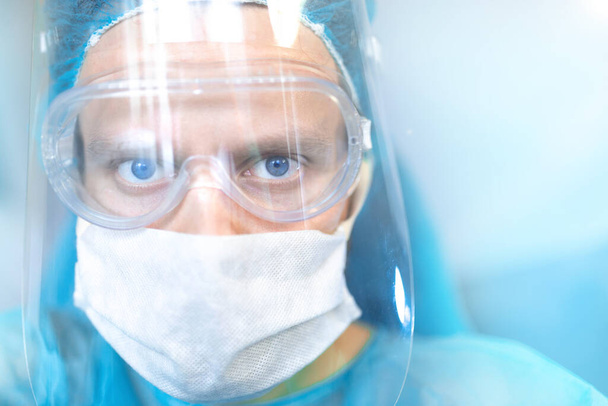 Gözlüklü, koruyucu kalkanlı ve maskeli bir cerrah. Genç uzman ameliyathanede çalışıyor. - Fotoğraf, Görsel