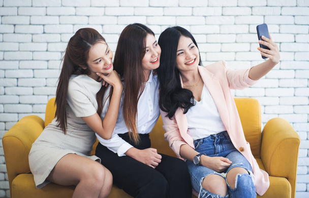 Счастливые воспоминания. Три молодые счастливые женщины друзья сидя на диване, глядя в сторону сделать селфи по телефону
. - Фото, изображение