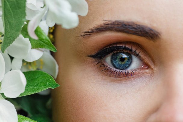 Beautiful white girl with flowers. A stunning brunette with a bouquet of white Apple trees near her eyes. Close-up of the face of a young beautiful woman with healthy clear skin - Photo, Image