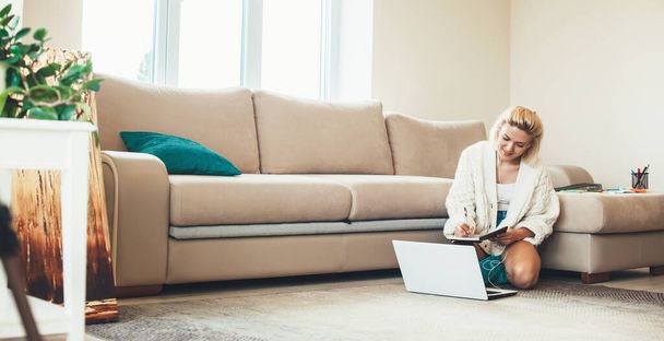Blondynka kaukaska kobieta siedzi na podłodze i robi notatki patrząc na laptopa - Zdjęcie, obraz