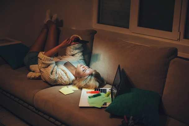 Unavený běloch student chatování na telefonu, zatímco sedí na pohovce po provedení domácích úkolů pozdě večer - Fotografie, Obrázek