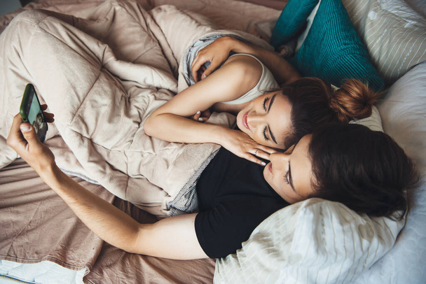 jong paar liggend in bed en omarmen terwijl glimlach en het maken van selfie bedekt met een quilt vroege ochtend - Foto, afbeelding