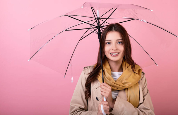 Portrait of beautiful young brunette lady wearing an autumn coat and scarf standing, smiling tenderly, isolated on a pink background, holding an umbrella, hiding from the rain, bad weather.Copy space. - Fotó, kép