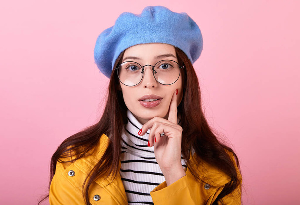 Sexy girl in a gentle blue beret, a striped blouse and a yellow rain jacket with glasses on a pink background. Lady holds her chin and thinks romantically looking to the right. Rainy day. Copy space. - Fotoğraf, Görsel