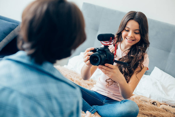 Cheerful beautiful girl sitting on the bed with a camera and smiling while recording a video of her friend - 写真・画像