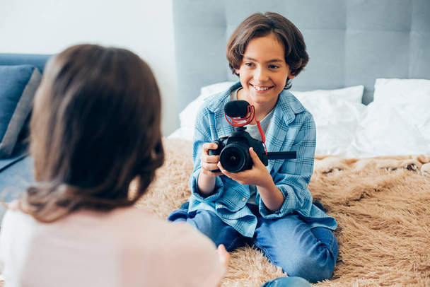 Positive boy sitting on the bed with a camera in his hand and smiling to a girl in front of him - Фото, изображение