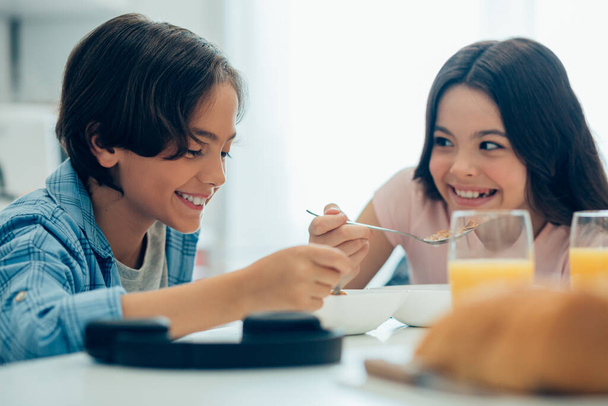 Pozitív barátságos fiú és lány ül az asztalnál egy pohár narancslével és mosolyog, miközben kukoricapelyhet eszik - Fotó, kép