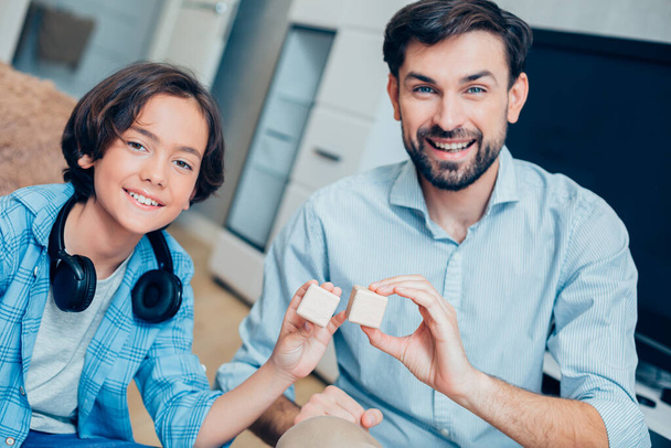 Happy young man and his smiling son sitting in the bedroom and showing two wooden pieces of the building set - Foto, Imagem