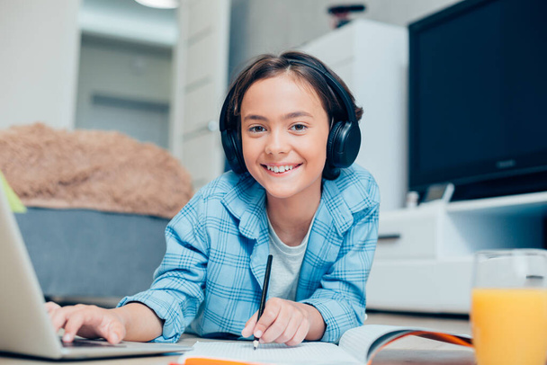 Positive boy wearing headphones and holding a pencil while lying on the floor in front of a laptop screen - Fotoğraf, Görsel
