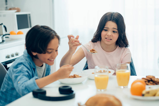 Onnellinen poika syö ja hymyilee siskonsa murjottaessa ja katsoessa häntä. Lusikallinen maissihiutaleita kädessään - Valokuva, kuva