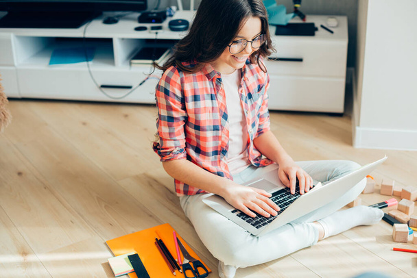 Contented girl in casual clothes and spectacles sitting on the floor with stationery supplies by her side and smiling while looking at the laptop screen - Фото, изображение