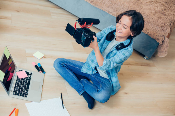 Gladsome child sitting on the floor with a camera and a microphone and making videos. Modern laptop and notes in front of him - 写真・画像