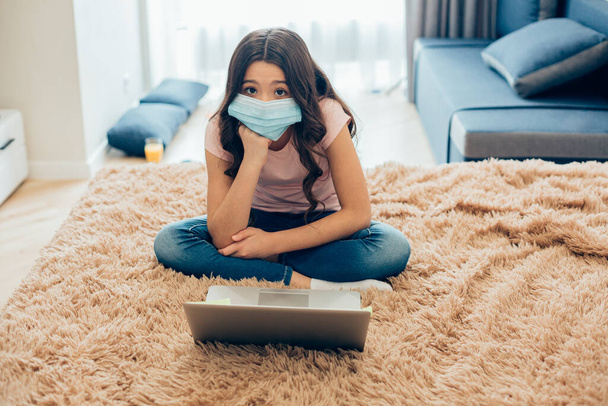 Tired long haired girl putting chin on her fist while sitting on the bed in front of a laptop. Medical mask on her face - Foto, imagen