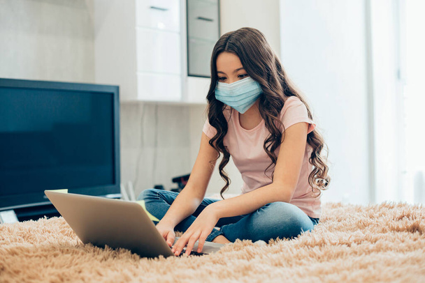 Long haired girl sitting on the bed with a mask on her face and looking at the screen of a modern laptop - Фото, изображение