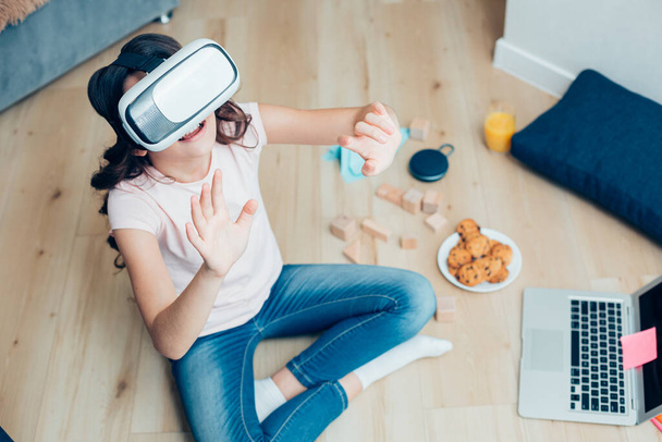 Child in casual clothes sitting on the floor with a laptop and a plate of cookies. Virtual reality device on her eyes - Photo, Image