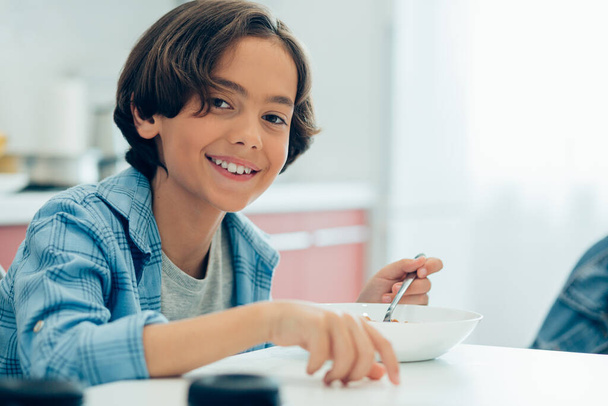 Positive smiling boy holding a spoon while sitting with a bowl of cornflakes at the kitchen table - Foto, Bild