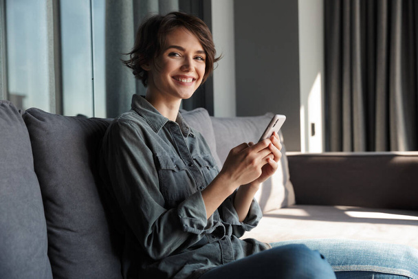 Image of beautiful young joyful woman using mobile phone and smiling while sitting on sofa at living room - Фото, зображення