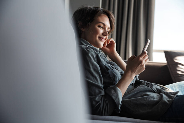Image of nice young happy woman using mobile phone and smiling while sitting on sofa at living room - Photo, Image