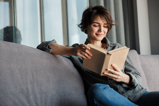 Image of brunette cute focused woman reading book and smiling while sitting on sofa at living room - Foto, Imagen