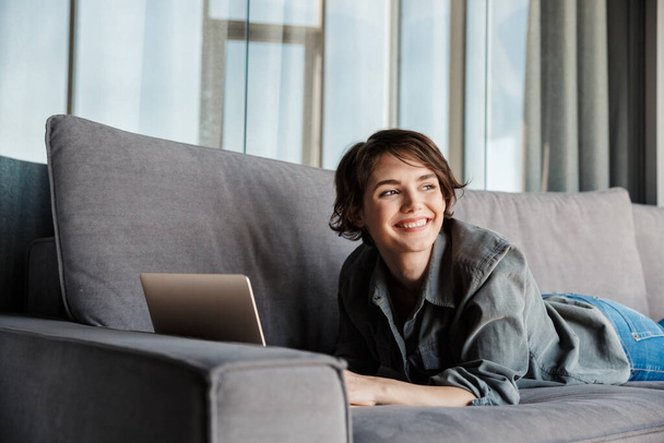 Image of caucasian young joyful woman using laptop and smiling while resting on sofa at living room - Φωτογραφία, εικόνα