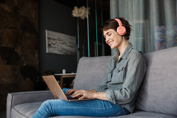 Image of nice young smiling woman using laptop and headphones while sitting on couch at living room - Foto, afbeelding