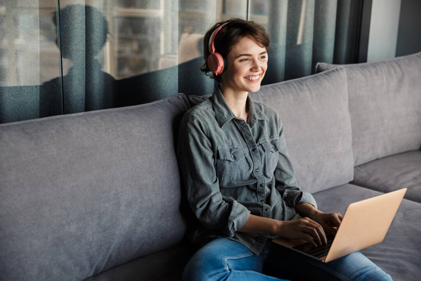 Image of nice young smiling woman using laptop and headphones while sitting on couch at living room - Foto, Imagem