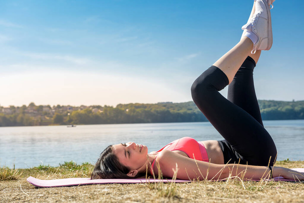 Die junge brünette Frau in Sportbekleidung streckt ihre Beine nach dem Training am See in der Nähe des Tages. Gesunder Lebensstil - Foto, Bild