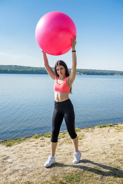   Jovem mulher flexível fazendo exercícios de fitness matinal na bola rosa perto do lago
 - Foto, Imagem