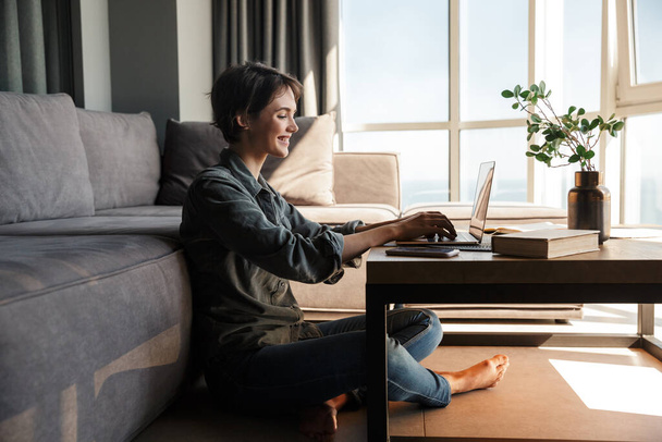 Image of nice young cheerful woman using laptop and smiling while sitting on floor at living room - Foto, imagen