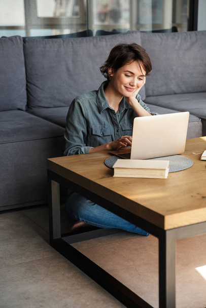 Image of nice young cheerful woman using laptop and smiling while sitting on floor at living room - Foto, Bild