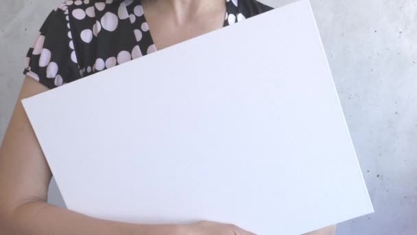 Γυναίκα που κρατάει στα χέρια της κενή λευκή αφίσα. Γκρι τοίχωμα στο φόντο - Πλάνα, βίντεο