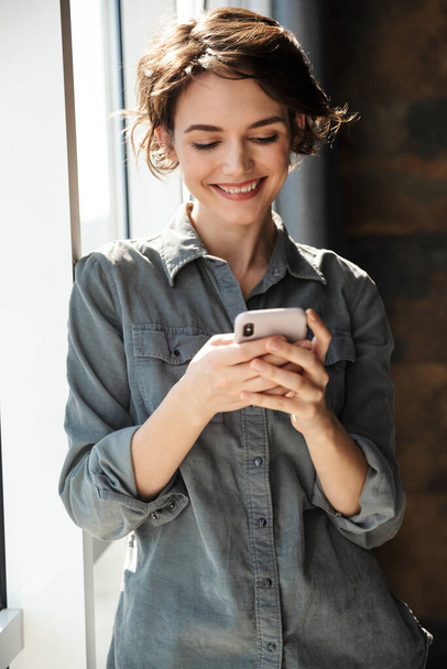 Image of beautiful young joyful woman using mobile phone and smiling while standing near window - Φωτογραφία, εικόνα