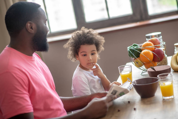 Dark-haired african american father reading a newspaper while having breakfast with his kid - Фото, изображение