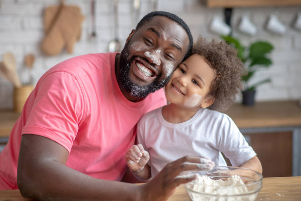 Dark-skinned father looking funny with face stained with flour, his daughter smiling - Fotoğraf, Görsel